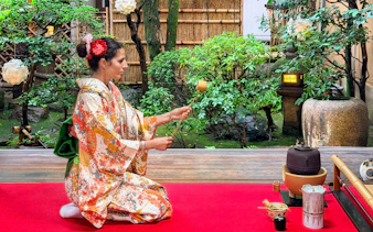 Person in a patterned kimono performing a Japanese tea ceremony in a garden setting.