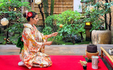 Person in a patterned kimono performing a Japanese tea ceremony in a garden setting.