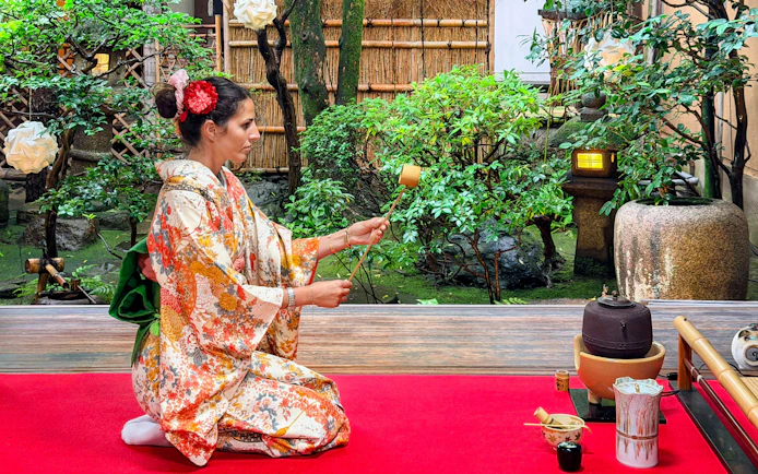 Person in a patterned kimono performing a Japanese tea ceremony in a garden setting.