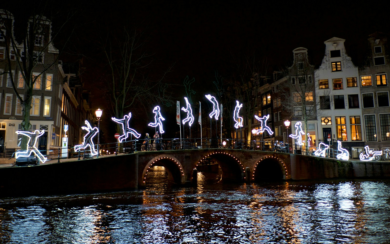 Illuminated figures on a canal bridge during Amsterdam Light Festival.