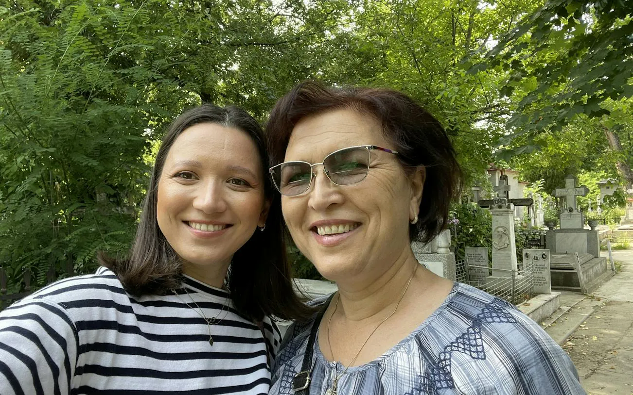 Two people smiling in Bellu Cemetery, Bucharest, surrounded by gravestones and greenery.