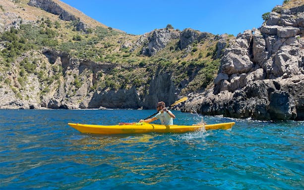 Kayaker paddling in clear blue waters at Ieranto Bay with rocky cliffs in the background.