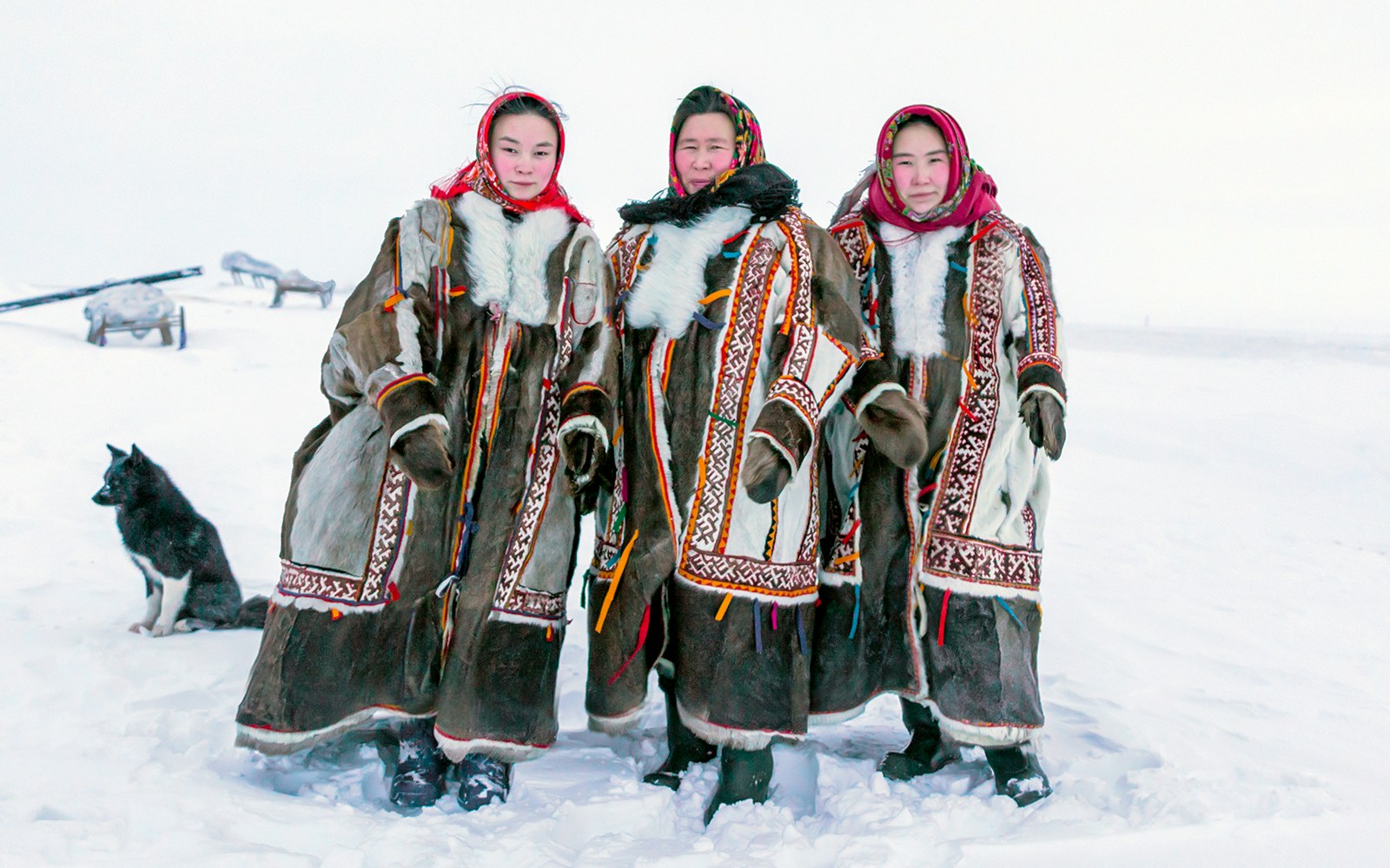 Three people in traditional fur clothing with a sled and dog in snowy Siberia.