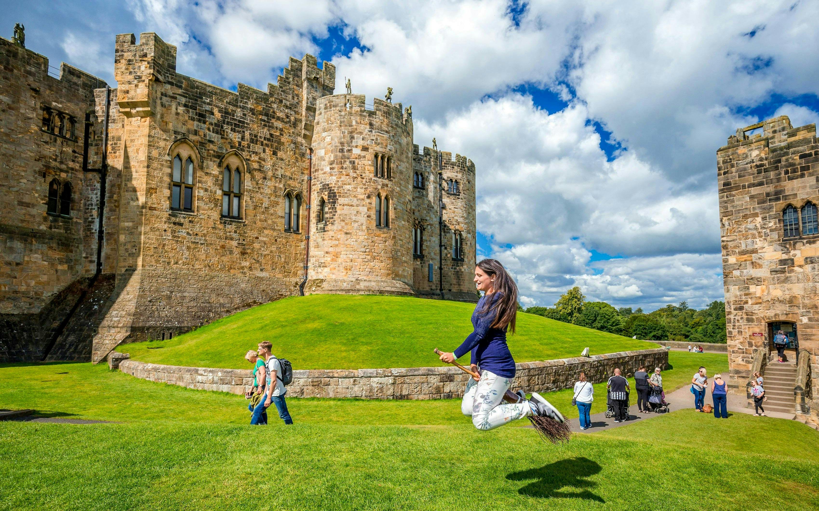 Person on broomstick in front of Alnwick Castle exterior, England.