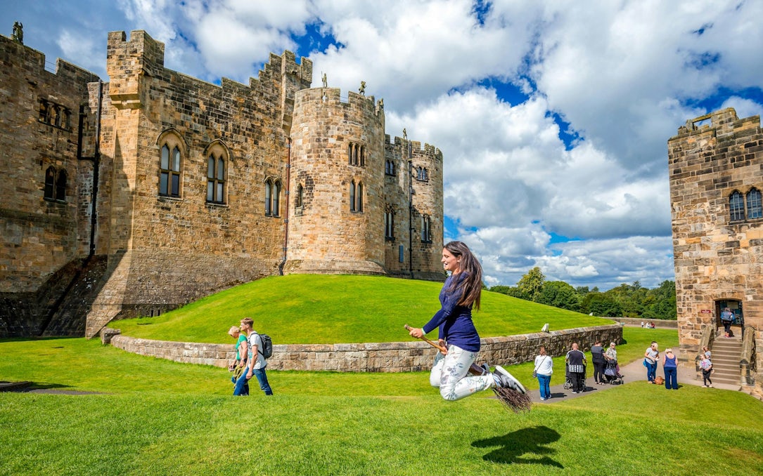 Person on broomstick in front of Alnwick Castle exterior, England.