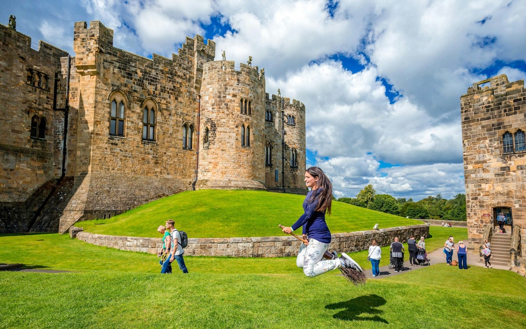Person on broomstick in front of Alnwick Castle exterior, England.