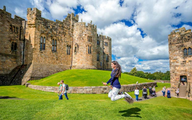 Person on broomstick in front of Alnwick Castle exterior, England.