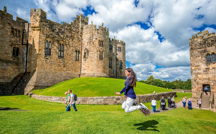 Person on broomstick in front of Alnwick Castle exterior, England.