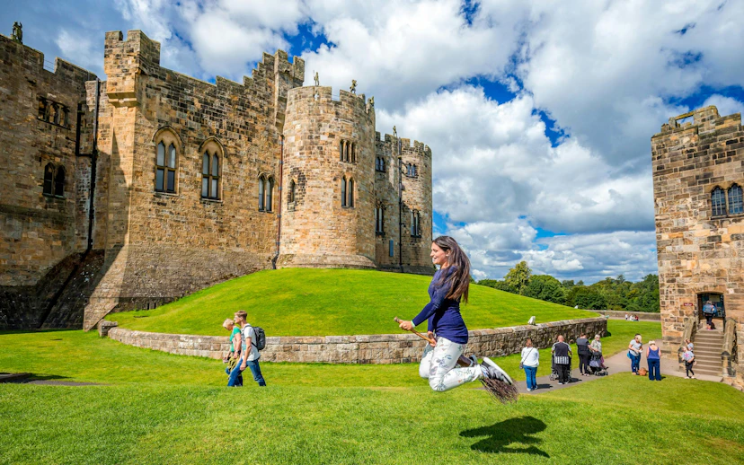 Person on broomstick in front of Alnwick Castle exterior, England.