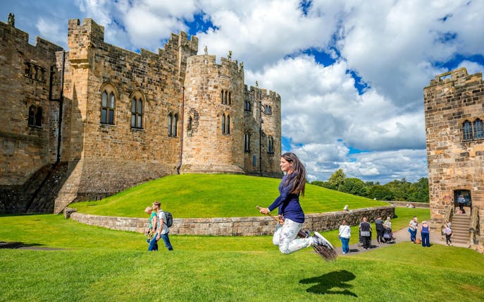 Person on broomstick in front of Alnwick Castle exterior, England.