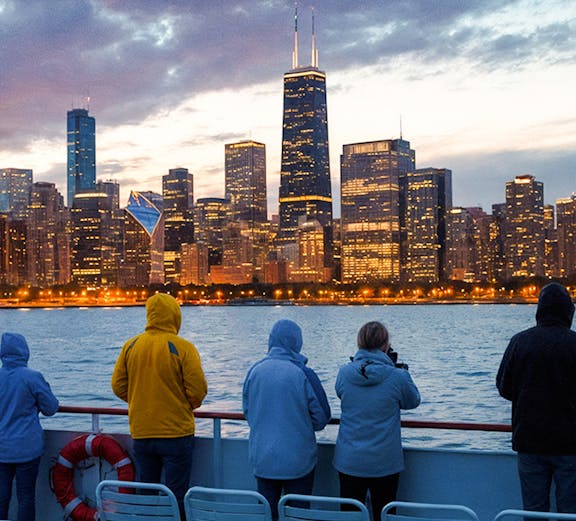People on a boat deck viewing Chicago skyline at dusk with water and cloudy sky.