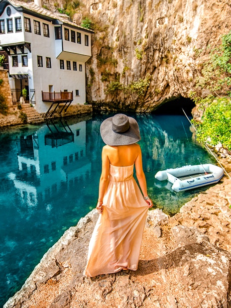 Guest overlooking Blagaj Buna river with historic house and cave in background.
