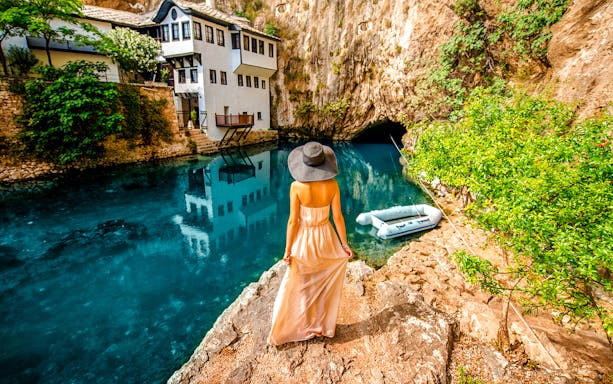 Guest overlooking Blagaj Buna river with historic house and cave in background.