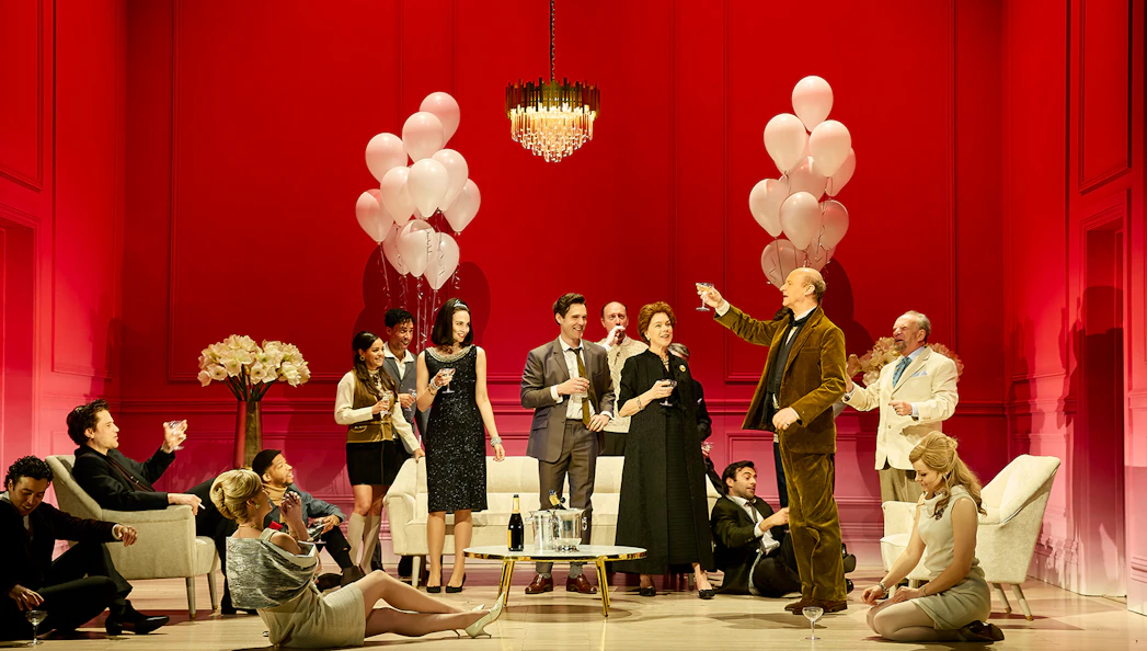 Group of people in formal attire celebrating with drinks and balloons in a red room.
