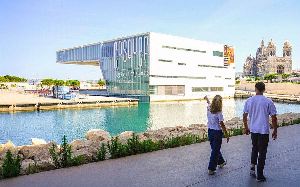 Cosquer Cave building in Marseille with people walking nearby, cathedral in background.