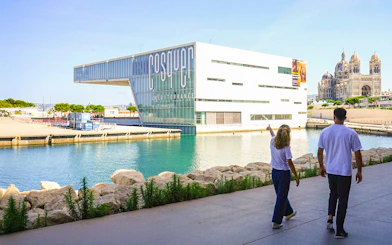 Cosquer Cave building in Marseille with people walking nearby, cathedral in background.