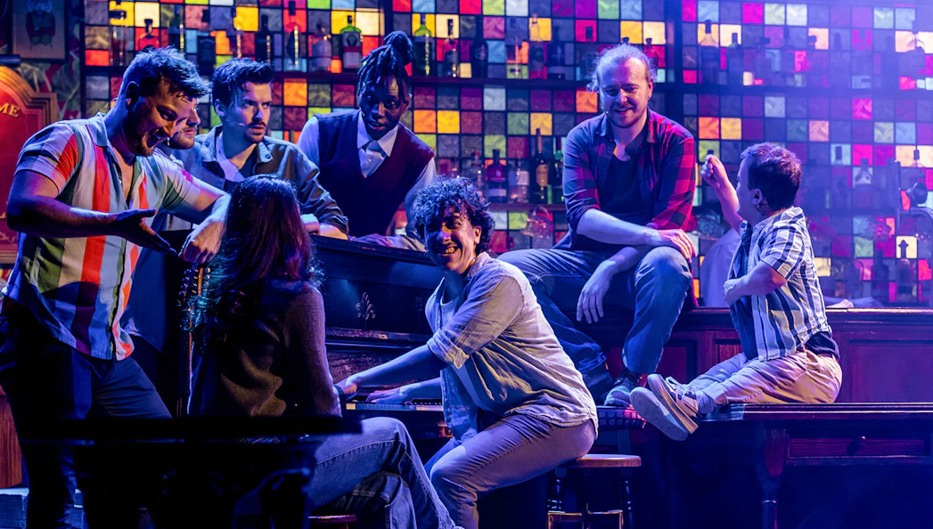 Performers from The Choir of Man show gathered around a piano on stage.
