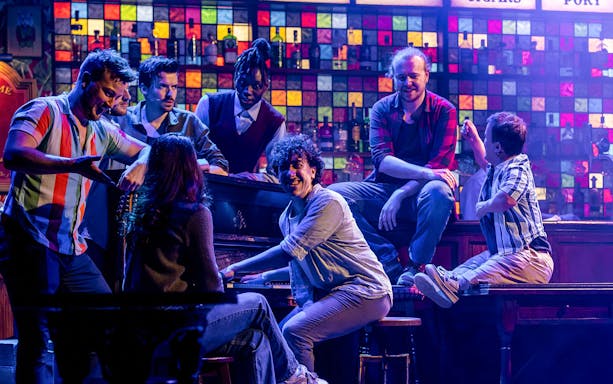 Performers from The Choir of Man show gathered around a piano on stage.