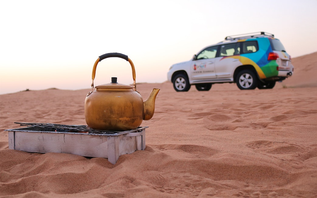 Tea brewing in a pot on sand during sunrise desert safari with a vehicle in the background.