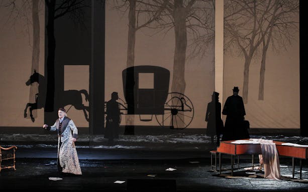 Opera scene from Les Contes d'Hoffmann with a performer on stage, shadowy carriage backdrop.