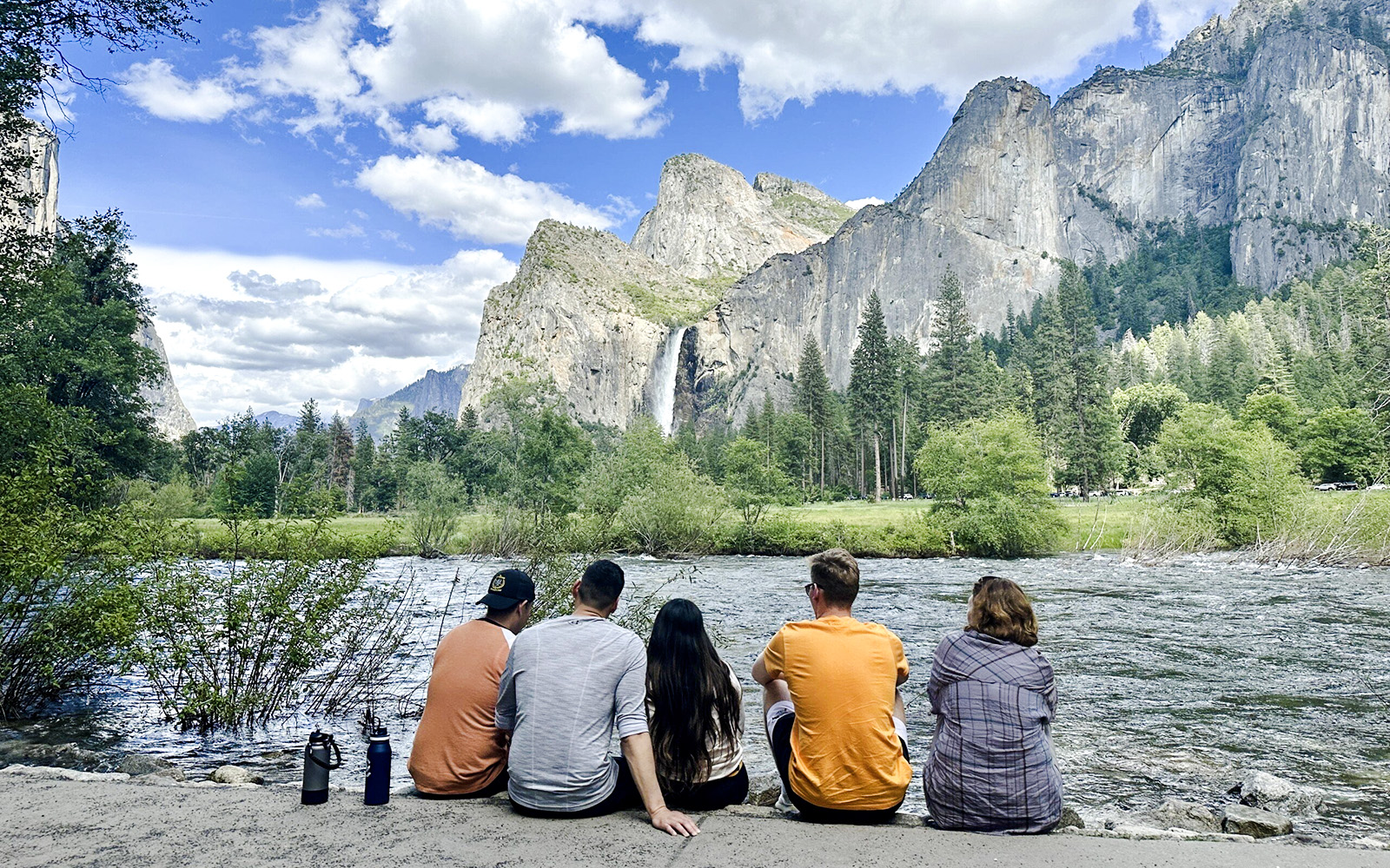 Guests sitting by a river with Yosemite Falls and cliffs in the background, Yosemite National Park.