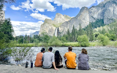 Guests sitting by a river with Yosemite Falls and cliffs in the background, Yosemite National Park.