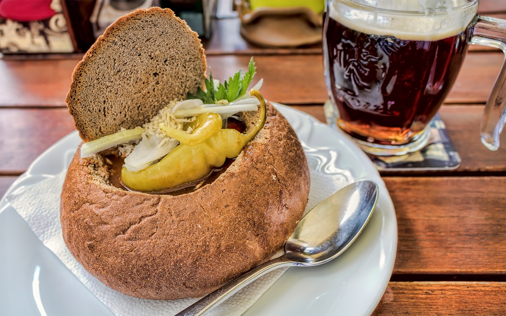 Bread bowl with beer goulash and pepper, accompanied by a mug of dark beer on a wooden table.