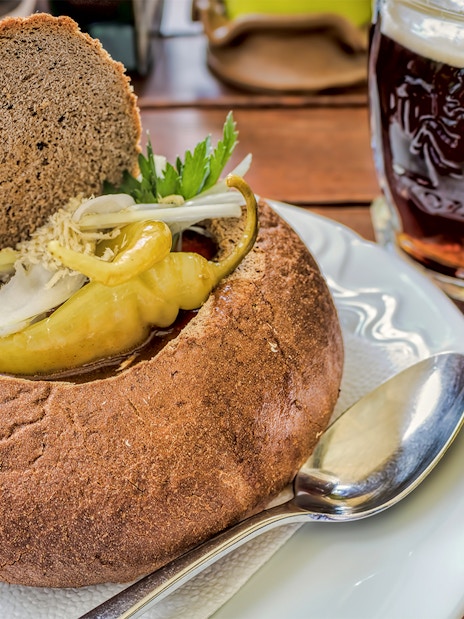 Bread bowl with beer goulash and pepper, accompanied by a mug of dark beer on a wooden table.