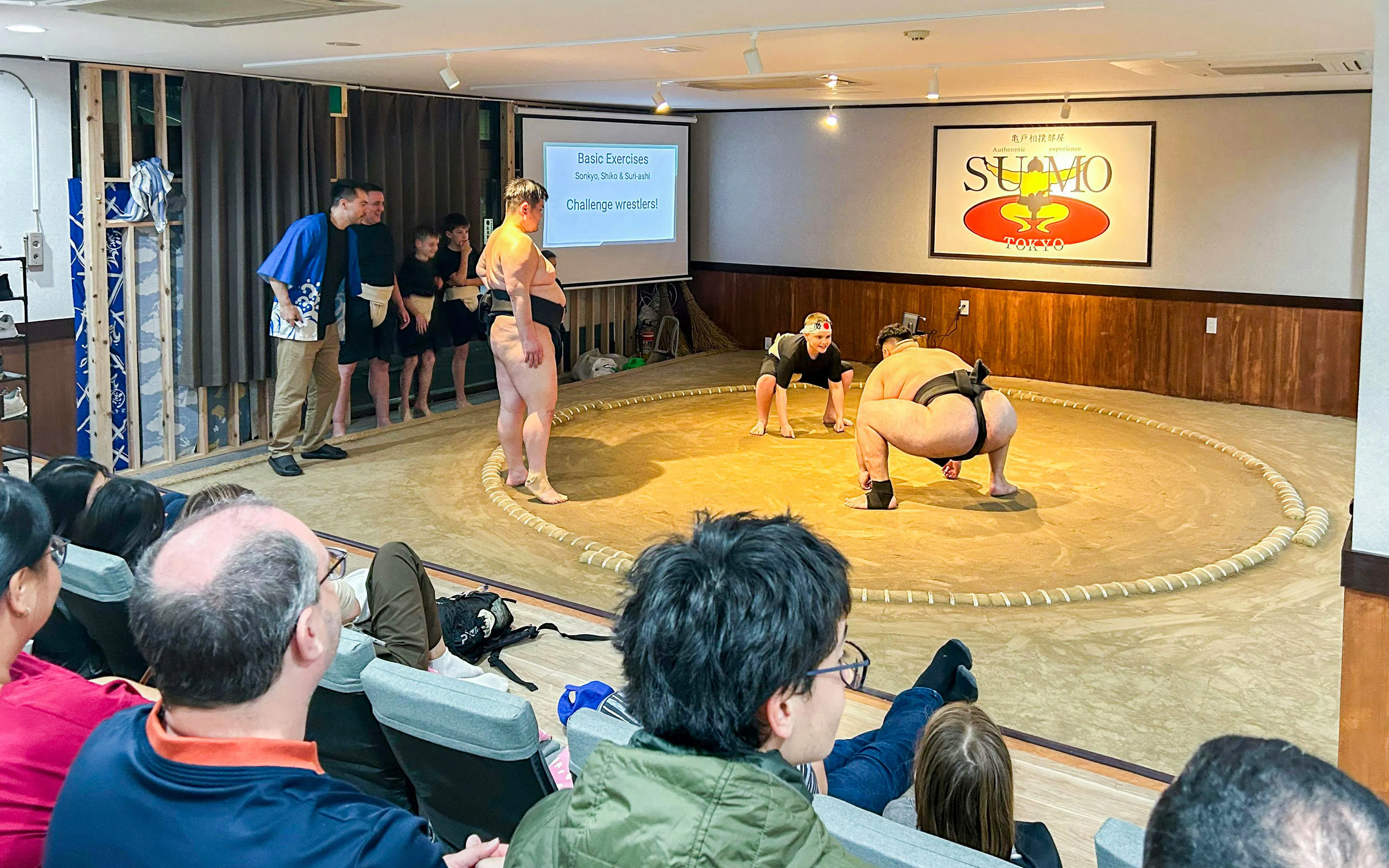 Sumo wrestlers demonstrating techniques in a Tokyo dojo with an audience watching.