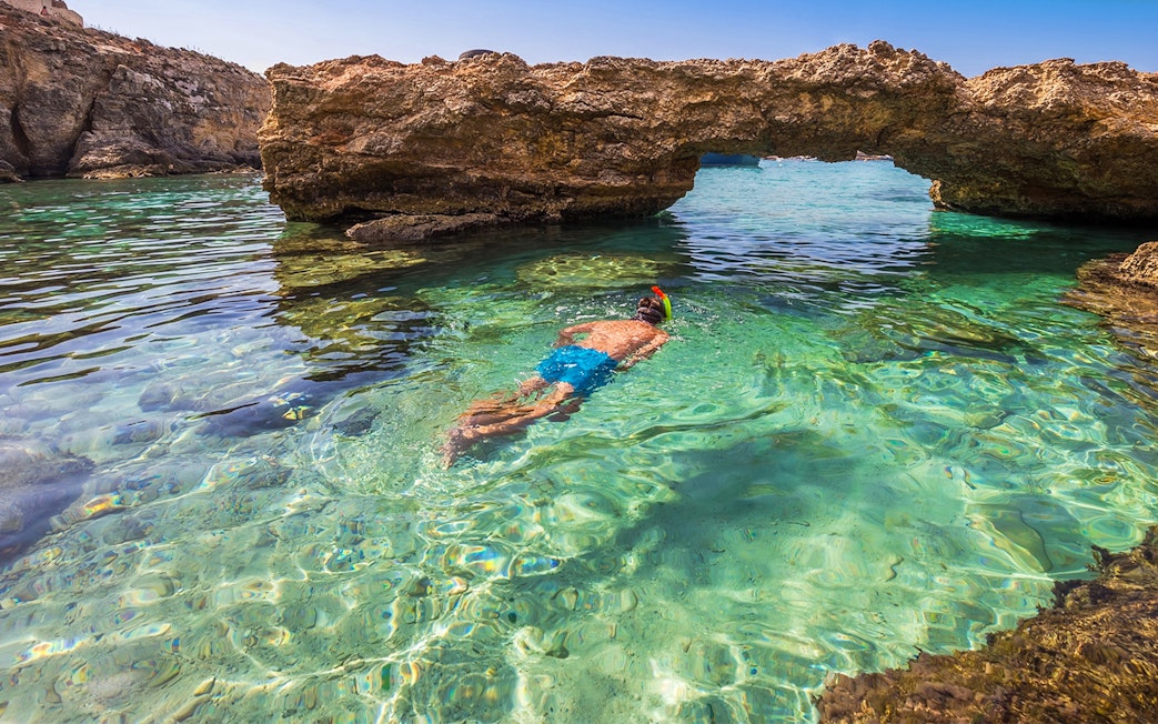 Snorkeler in clear waters near rock arch in Comino, Malta.