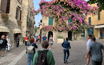 Tourists walking in a cobblestone street in Sirmione, Italy, with a stone building covered in vibrant flowers.