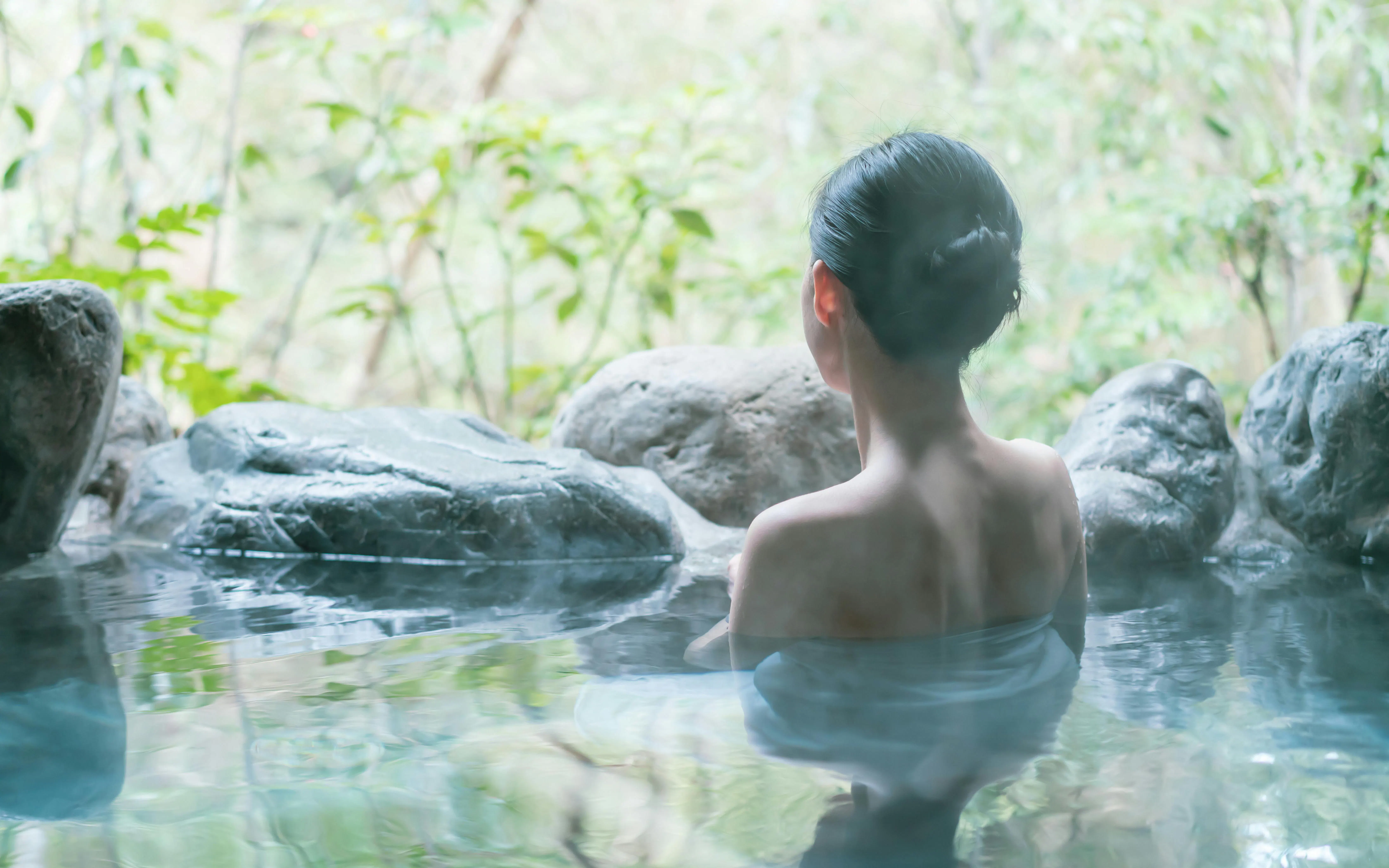 Woman relaxing in a natural hot spring onsen surrounded by rocks and greenery.