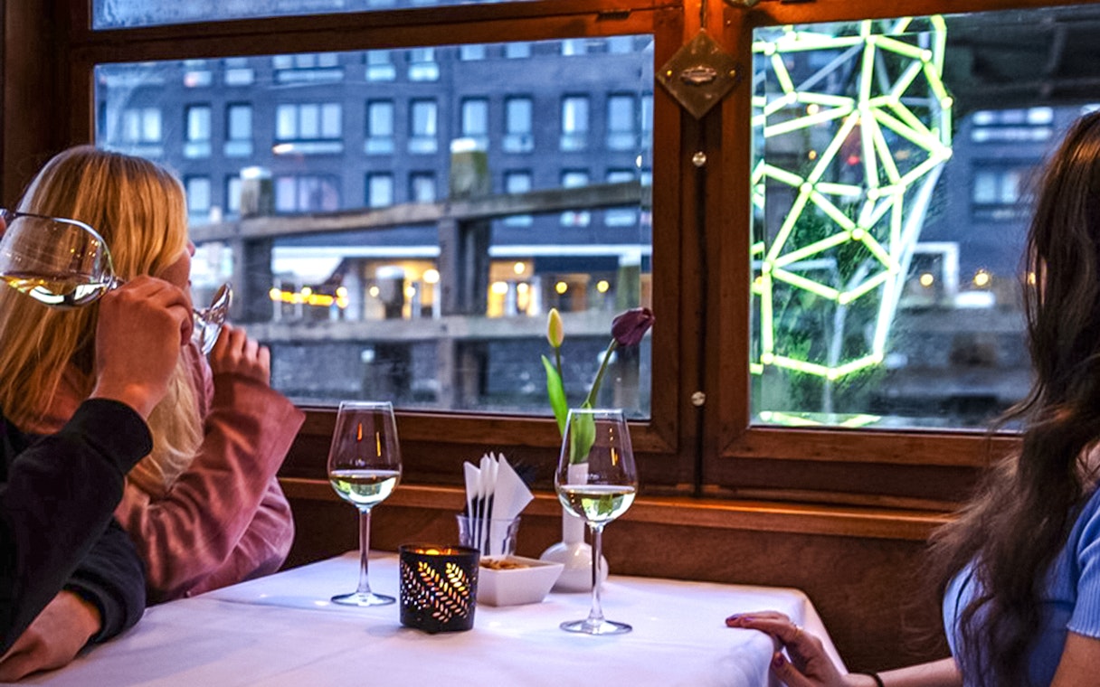 Guests enjoying wine on an Amsterdam Light Festival cruise with illuminated art outside.