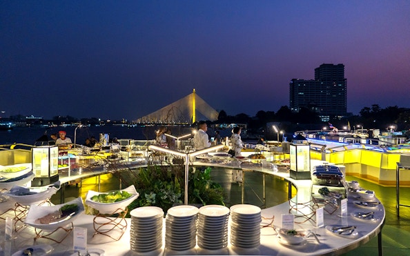 Buffet dinner setup on Chao Phraya cruise with illuminated bridge in Bangkok.