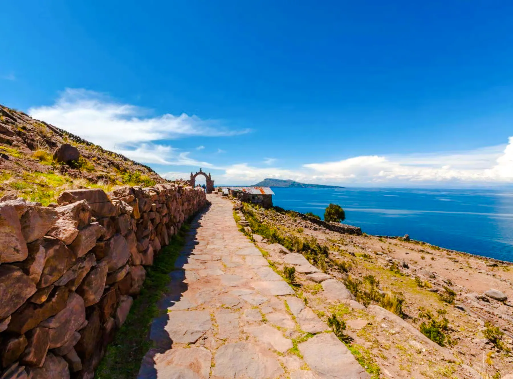 Stone path leading to archway on Taquile Island, Lake Titicaca, with distant mountains and blue sky.