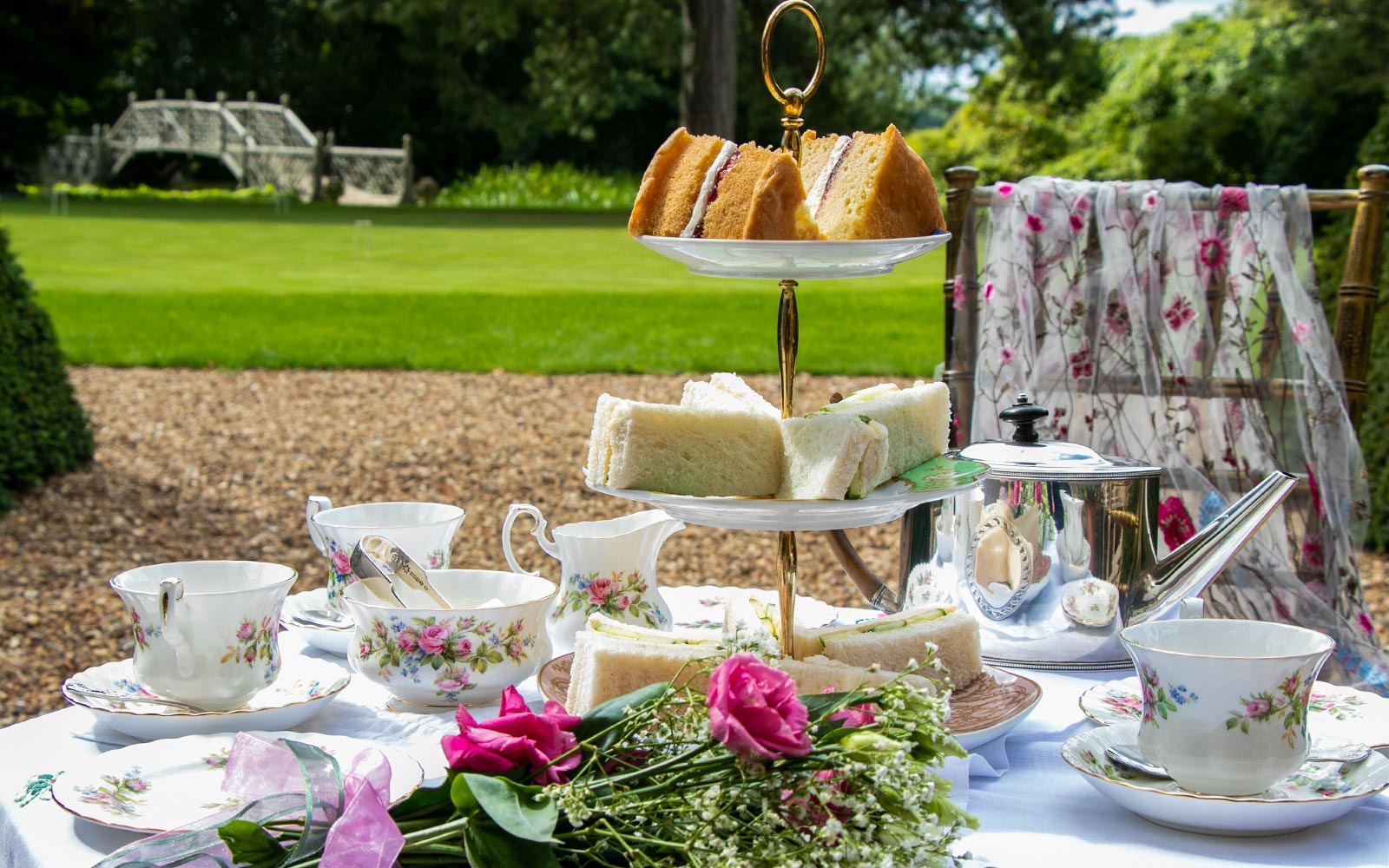Afternoon tea setup with tiered sandwich platter and floral teacups in a garden setting.