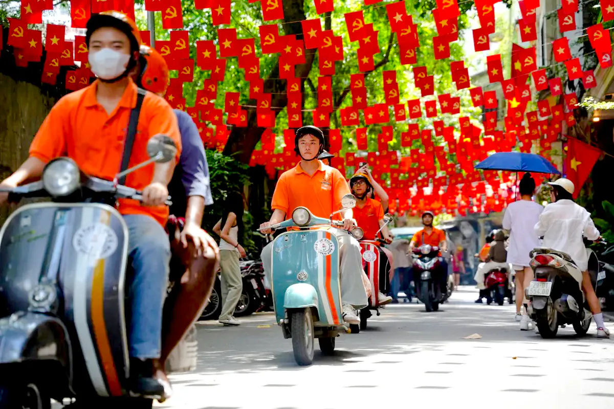 Vespa riders on a street in Hanoi with red flags overhead during a food adventure tour.