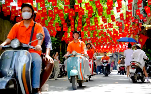 Vespa riders on a street in Hanoi with red flags overhead during a food adventure tour.