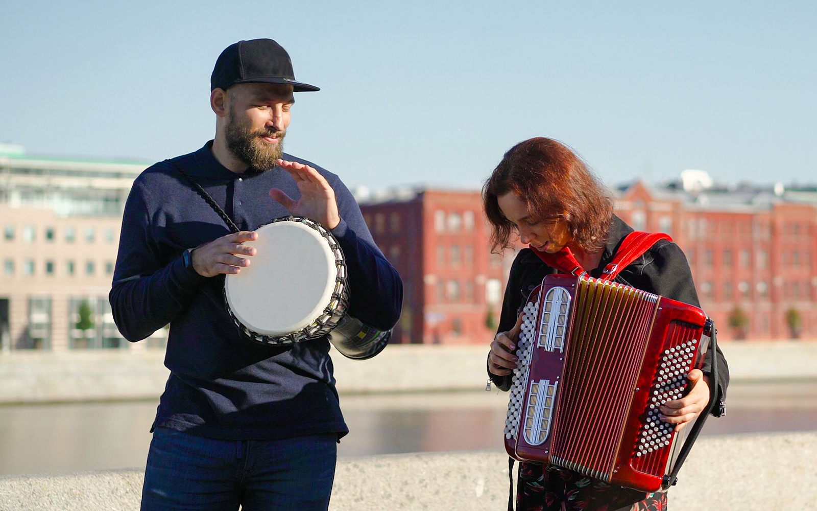 Street musicians playing drum and accordion by a riverside in an urban setting.