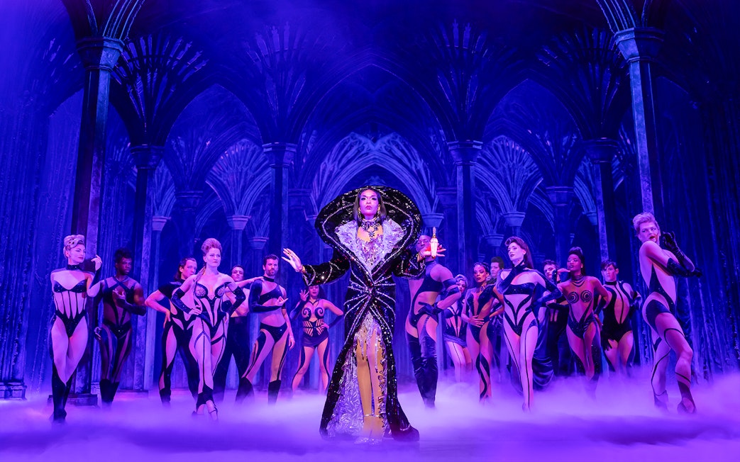 Performer in elaborate costume with dancers on stage in "Death Becomes Her" show.