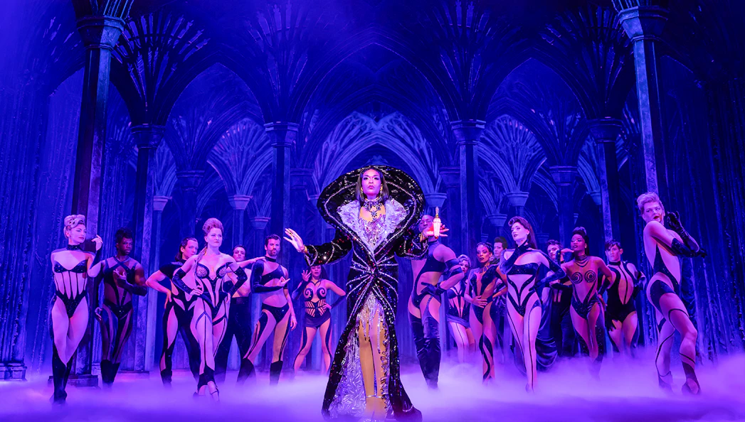 Performer in elaborate costume with dancers on stage in "Death Becomes Her" show.