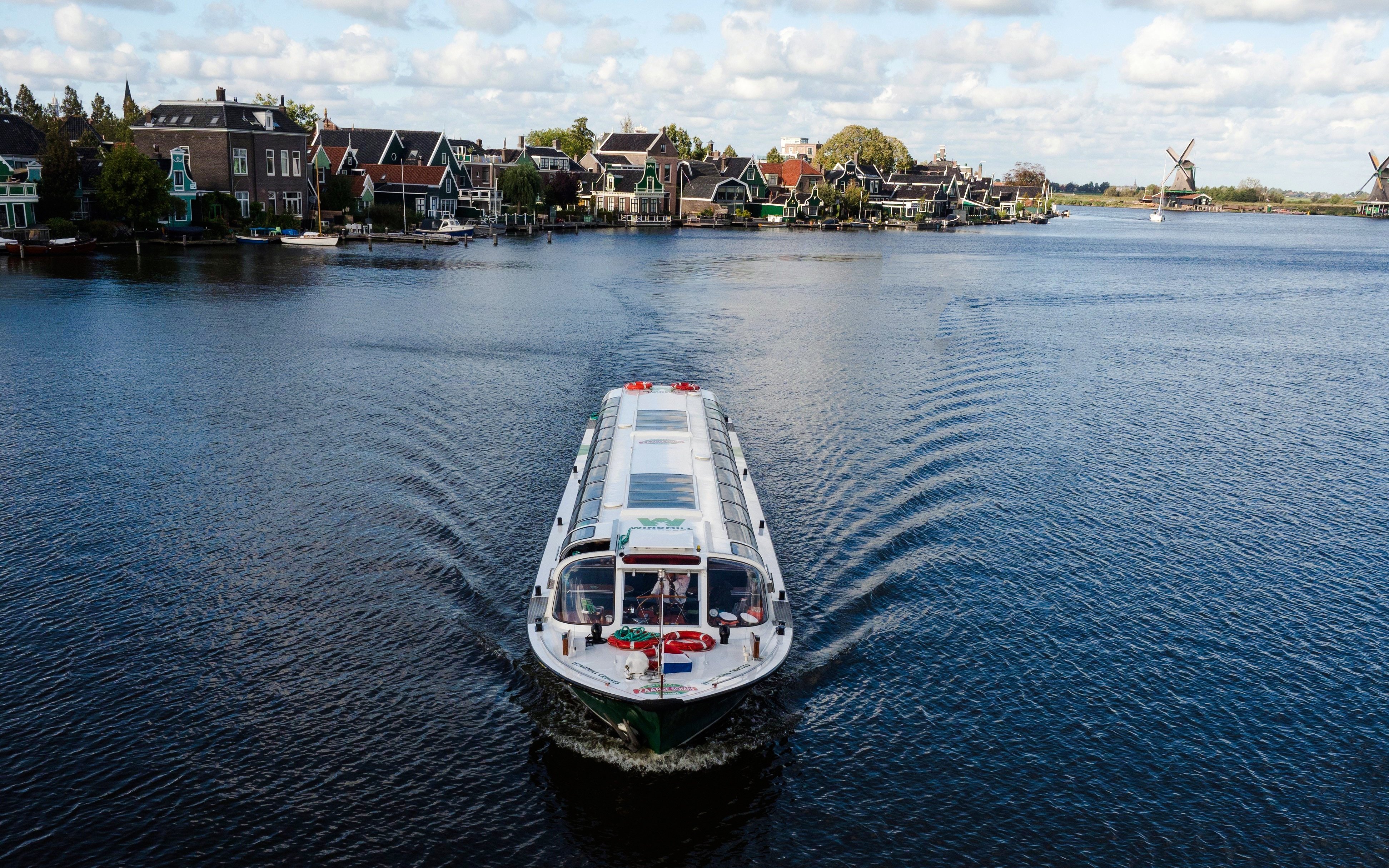 Cruise boat on a canal in Amsterdam with windmills and traditional houses in the background.