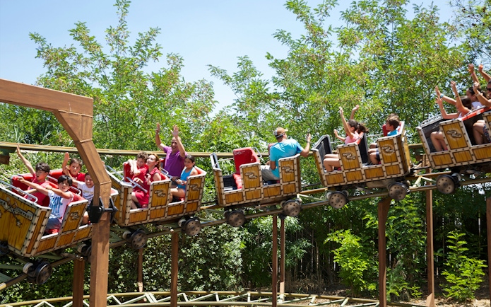Roller coaster ride with passengers at Isla Magica, Seville, surrounded by greenery.