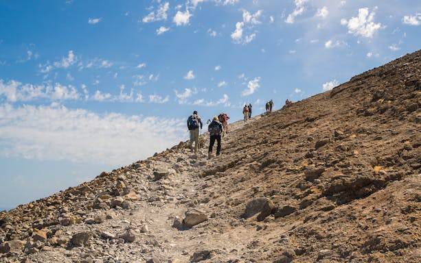 Hikers ascending rocky path on Vulcano Island under blue sky.