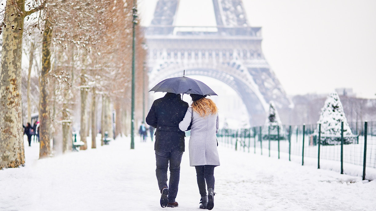 Couple walking under umbrella near Eiffel Tower in snowy Paris winter.