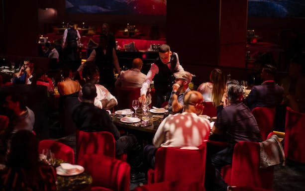 Dining experience at Paris Paradis with guests seated at red velvet chairs and a waiter serving wine.