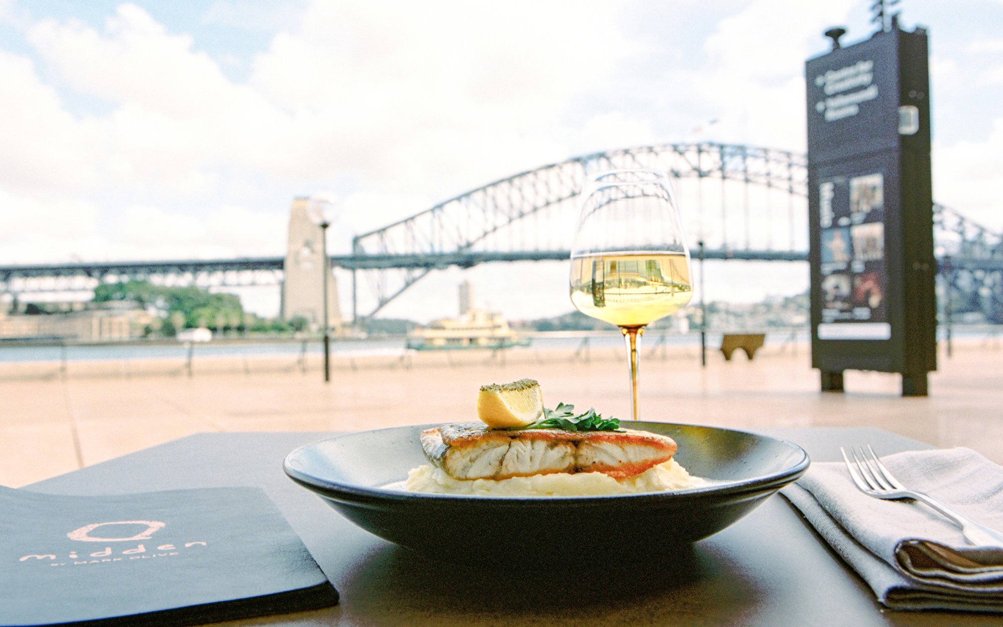 Grilled fish dish at Midden by Mark Olive, Sydney Opera House, with Harbour Bridge view.