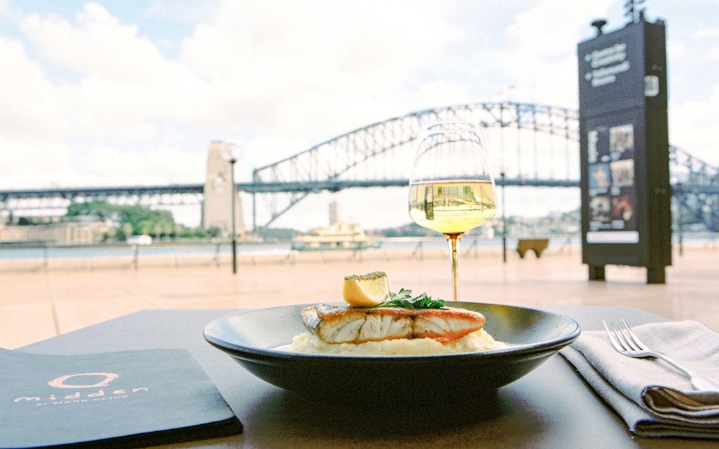 Grilled fish dish at Midden by Mark Olive, Sydney Opera House, with Harbour Bridge view.