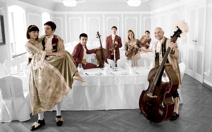 Musicians in period costumes with instruments at a dining table in Salzburg concert setting.