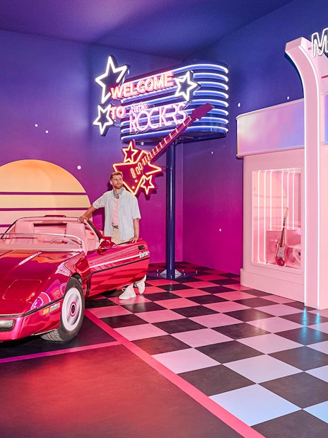 Ken at Barbie The Dream Experience in Amsterdam with a red convertible and neon signs.
